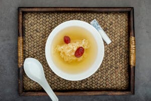 Top-down view of a white bowl containing traditional tonic with red dates on a woven wooden tray, illustrating bird's nest soup benefits.