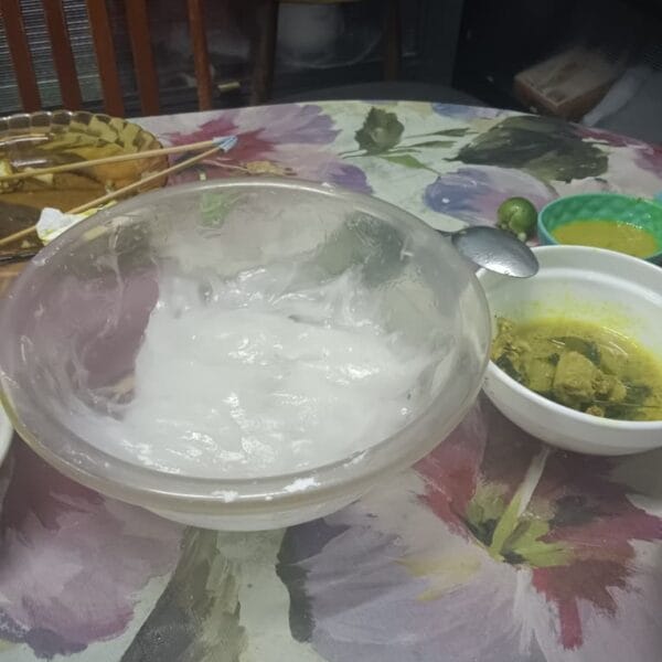A glass bowl filled with sticky white ambuyat, a traditional Borneo sago delicacy, served with a side of yellow turmeric curry on a floral tablecloth.