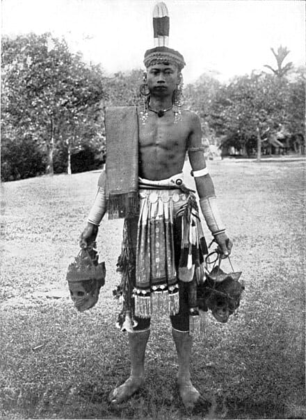 A vintage black-and-white photograph of a tribal warrior in ceremonial dress holding two trophy skulls, illustrating the fierce history of headhunting in Borneo and the indigenous cultures encountered by Z Special Unit during WWII.