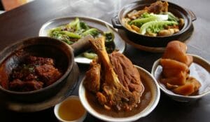 A table spread featuring authentic bak kut teh malaysia dishes, including a large "Big Bone" (Tua Kut) in herbal soup, a clay pot of savory dry bak kut teh, braised trotters, stir-fried vegetables, and Chinese tea.