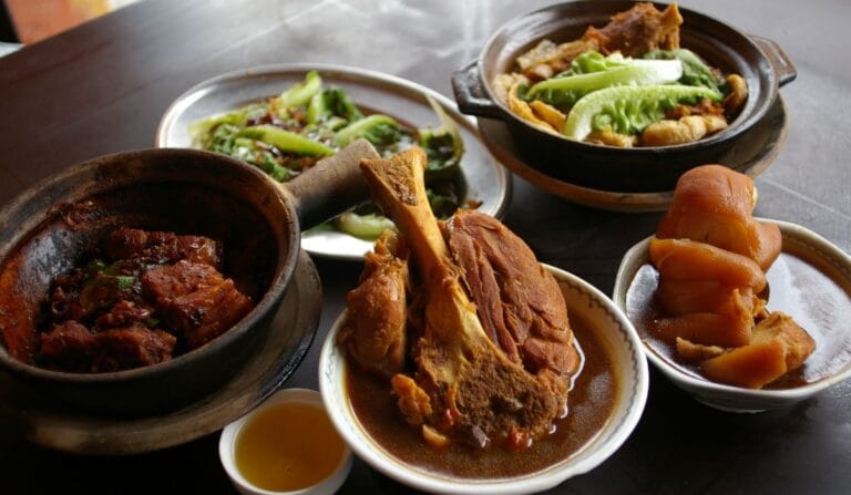 A table spread featuring authentic bak kut teh malaysia dishes, including a large "Big Bone" (Tua Kut) in herbal soup, a clay pot of savory dry bak kut teh, braised trotters, stir-fried vegetables, and Chinese tea.