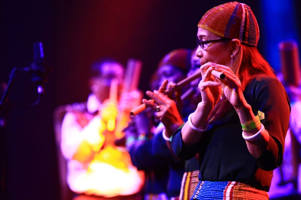 Close-up side profile of a woman playing a bamboo flute while wearing a traditional beaded headband.