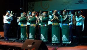 A large bamboo music ensemble performing on stage, featuring musicians in traditional green Kadazan-Dusun attire playing bamboo flutes and large bamboo bass pipes.