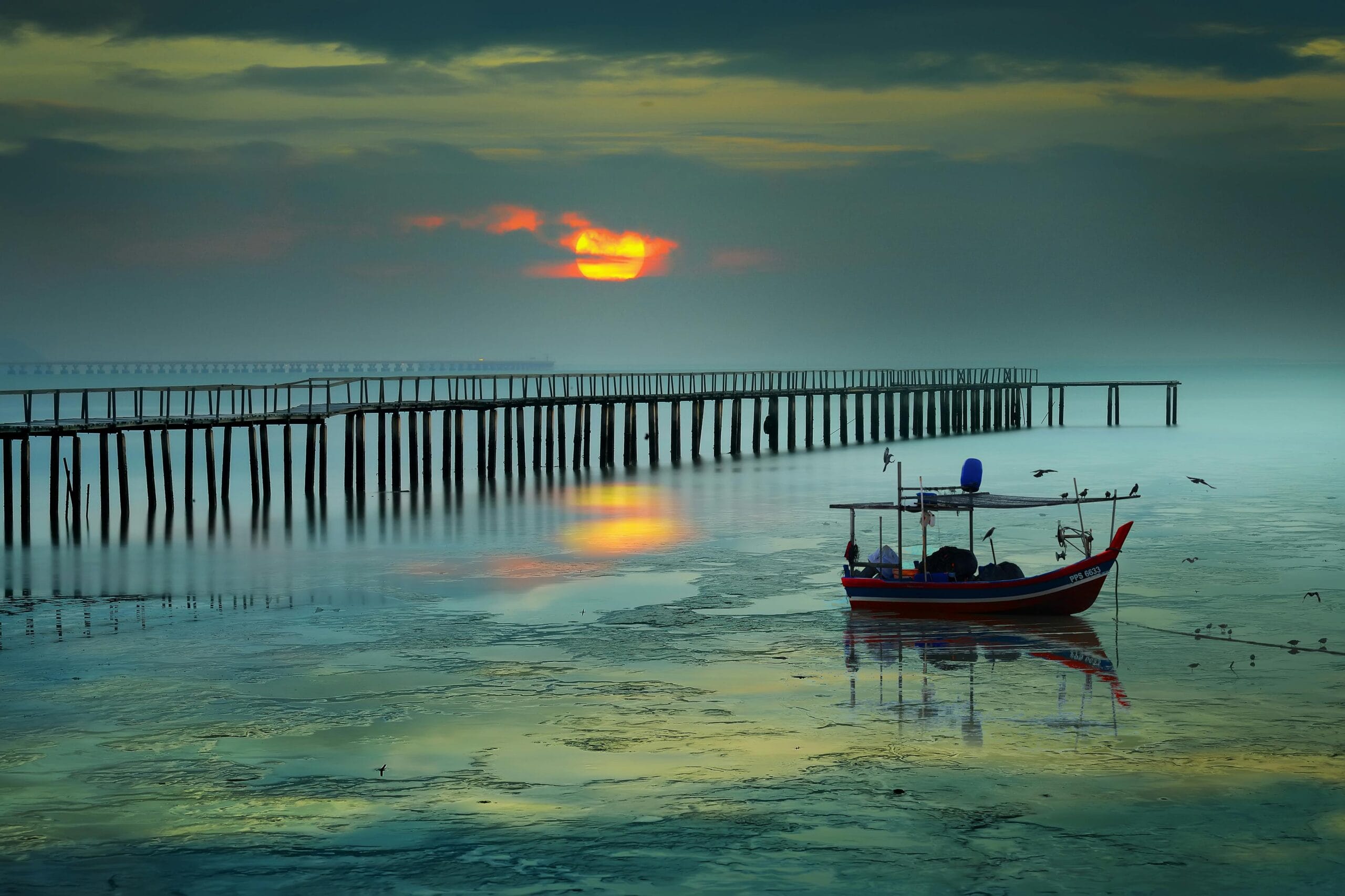 A dramatic silhouette of a long wooden pier and a small fishing boat floating on calm waters during sunset at the Penang jetty chew.
