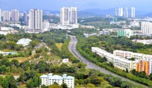 High-angle aerial view of Cyberjaya, Malaysia, featuring modern high-rise buildings, colorful residential blocks, and a winding highway surrounded by lush green trees under a soft, overcast sky.