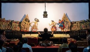 A traditional Fusion Wayang Kulit performance featuring a master puppeteer manipulating intricate leather puppets alongside modern superhero characters like Superman and Batman against a backlit screen.