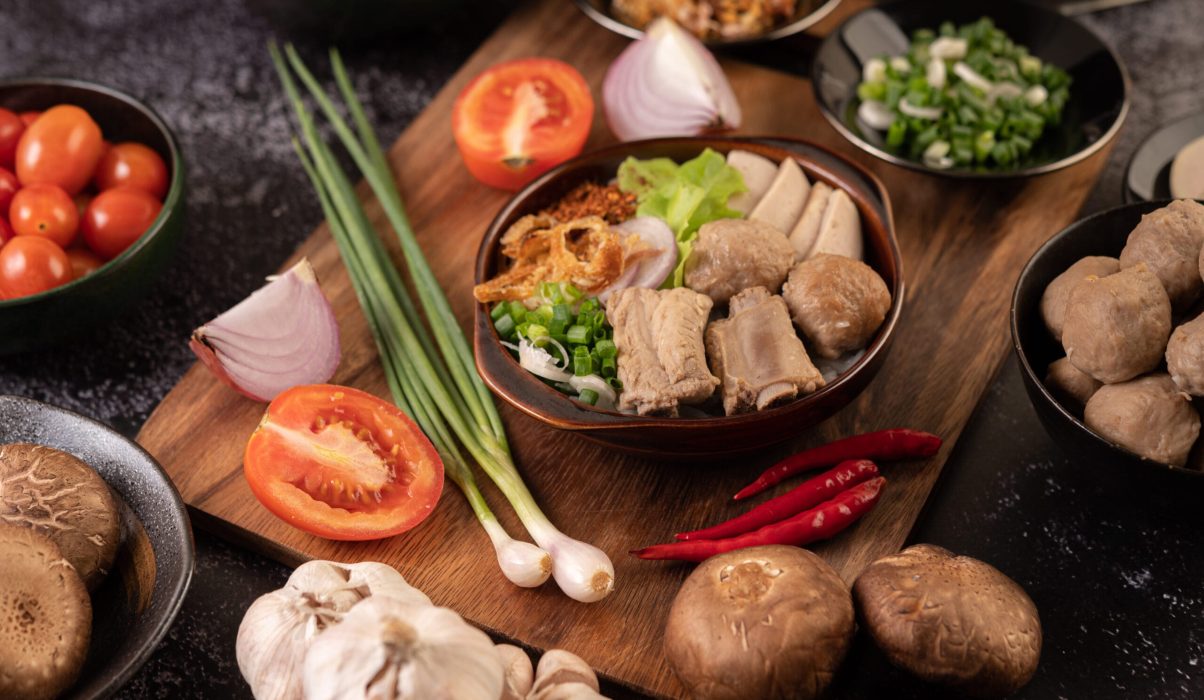 A rustic wooden board displaying fresh ingredients for a bak kut teh Malaysia recipe, including pork ribs, whole garlic bulbs, shiitake mushrooms, and fresh chilies.