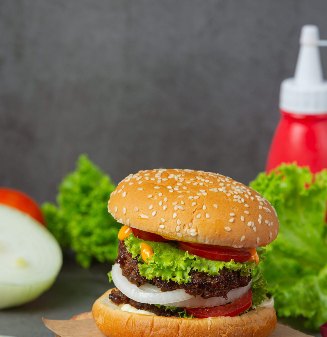 A vertical close-up of a juicy burger in Malaysia featuring a thick meat patty, white onion rings, fresh lettuce, and tomatoes next to a ketchup bottle.