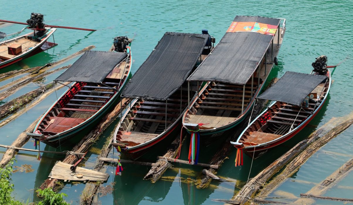 Traditional wooden longboats with black canopies and colorful ribbons docked at the river jetty in Kuala Tahan, used for transport to Taman Negara National Park.